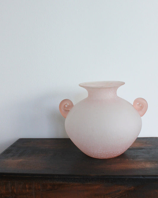 Pink glass vase with round handles on a wooden surface against a white wall