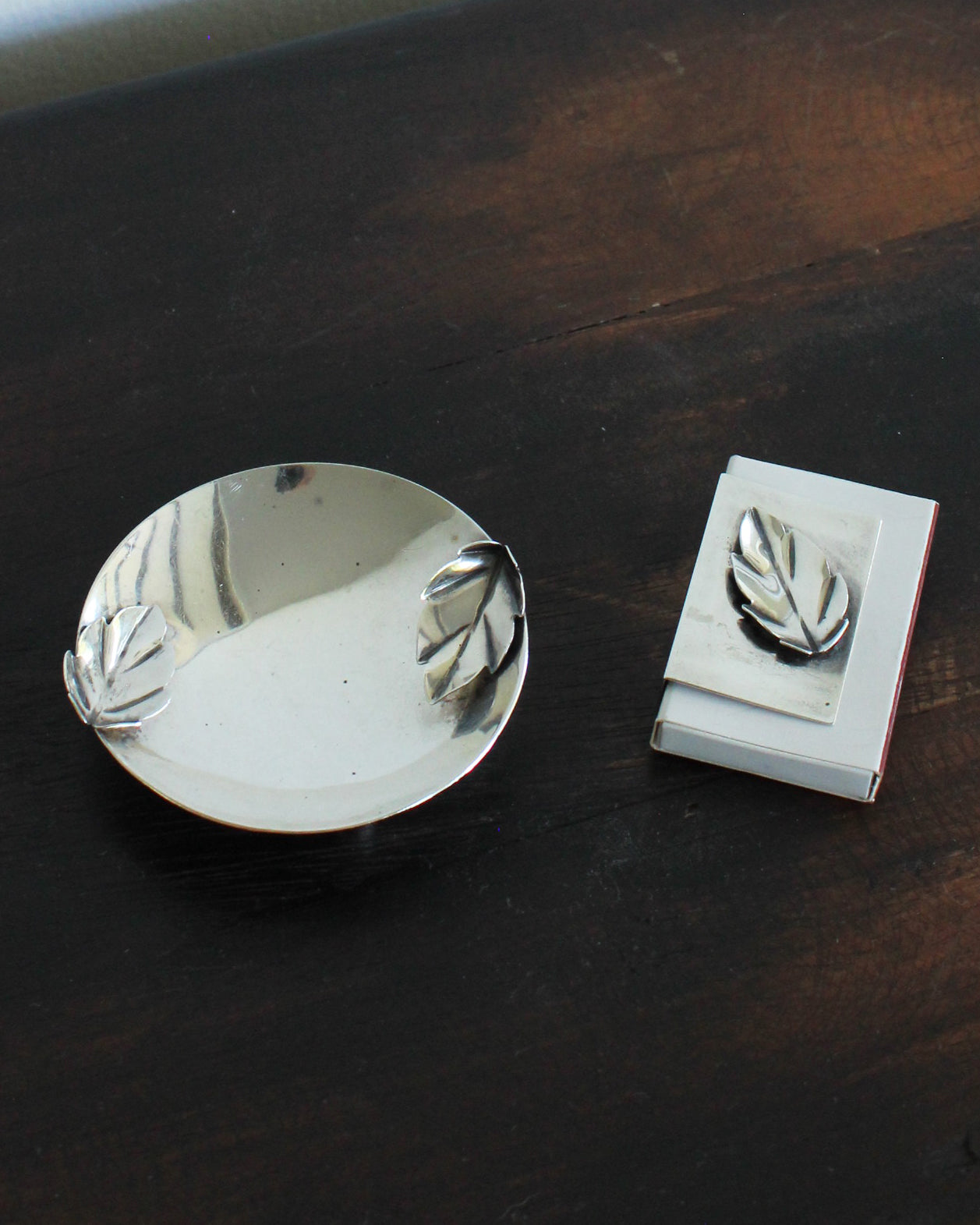 Metallic leaf-patterned ashtray and matchbook holder on a dark wooden surface