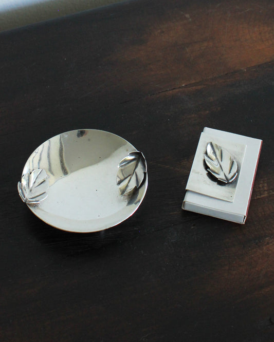 Metallic leaf-patterned ashtray and matchbook holder on a dark wooden surface